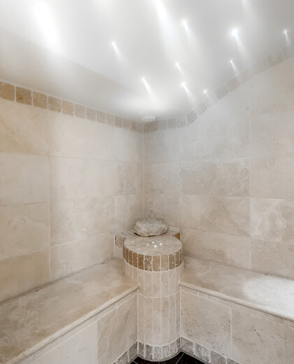 Ein modernes Dampfbad mit Wänden und Bänken aus beigen Steinfliesen, einem kleinen gefliesten Springbrunnen in der Mitte und Einbauleuchten an der Decke, die eine entspannte Atmosphäre schaffen.