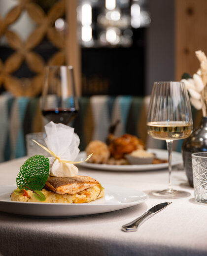 A close-up of a restaurant table featuring two plates of gourmet dishes, a glass of red wine and a glass of white wine, with an elegant interior in the background.
