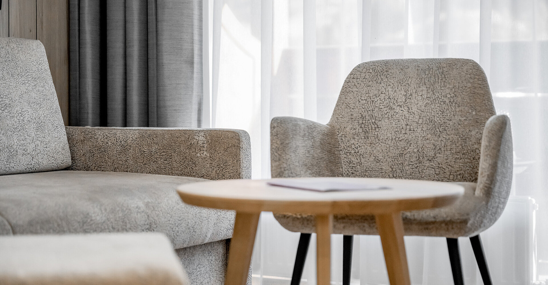A modern living room with a beige sofa, a matching armchair and a round wooden coffee table in front of white, sheer curtains.