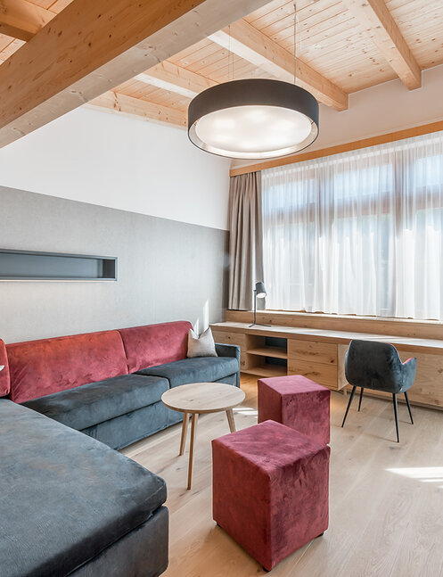 Modern living room with large windows, a gray and red corner sofa, two red poufs, a round coffee table, wooden floors, a desk with a chair and a ceiling with exposed wooden beams.