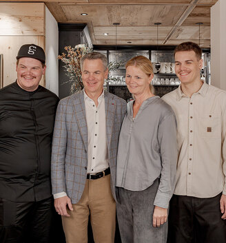 Four people are standing in a room, smiling at the camera. One of them is wearing a black outfit and a cap, while the others are dressed in light-colored casual and business attire. Wood paneling and a painting featuring a mountain scene are in the background.
