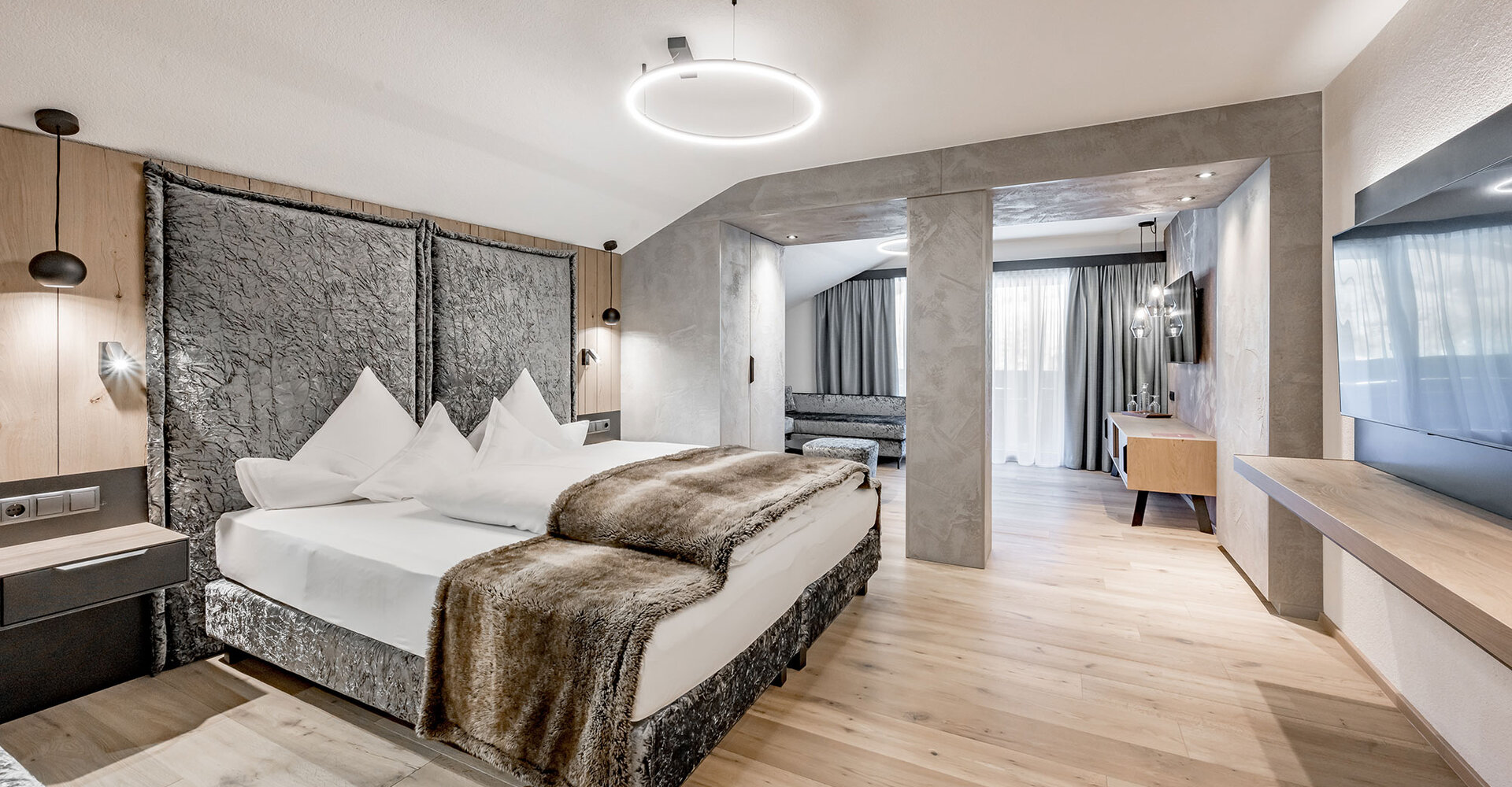 Modern hotel room featuring a large bed, décor in shades of gray and brown, a wall-mounted TV, wooden floors and a separate seating area by the window with sheer curtains that let in natural light.