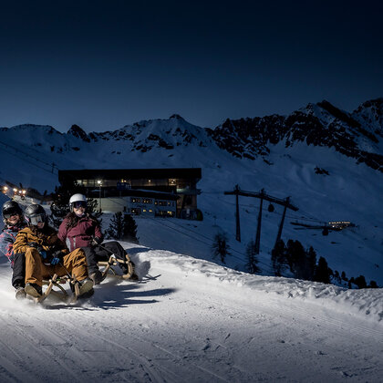 Three people wearing helmets and winter clothing are speeding down a snowy mountain slope at night with a ski hut and mountains in the background.