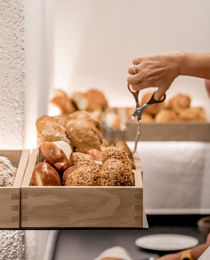 A person uses tongs to take bread from a wooden tray that is sitting on a buffet counter and is filled with various bread rolls.