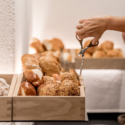 Eine Person nimmt mit einer Zange Brot von einem Holztablett, das auf einer Buffet-Theke steht und mit verschiedenen Brötchen gefüllt ist.