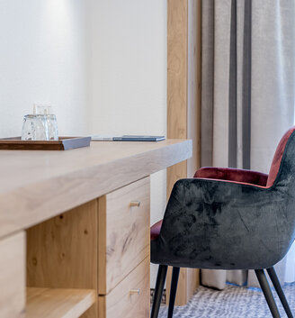 A modern desk with wooden drawers, a glass of water on a tray and a padded gray chair next to a window with sheer curtains.