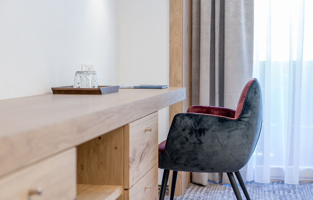 Ein moderner Schreibtisch mit Holzschubladen, ein Glas Wasser auf einem Tablett und ein gepolsterter grauer Stuhl, neben einem Fenster mit transparenten Vorhängen.