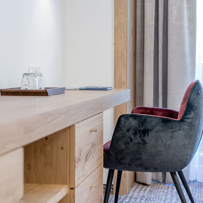 Ein moderner Schreibtisch mit Holzschubladen, ein Glas Wasser auf einem Tablett und ein gepolsterter grauer Stuhl, neben einem Fenster mit transparenten Vorhängen.