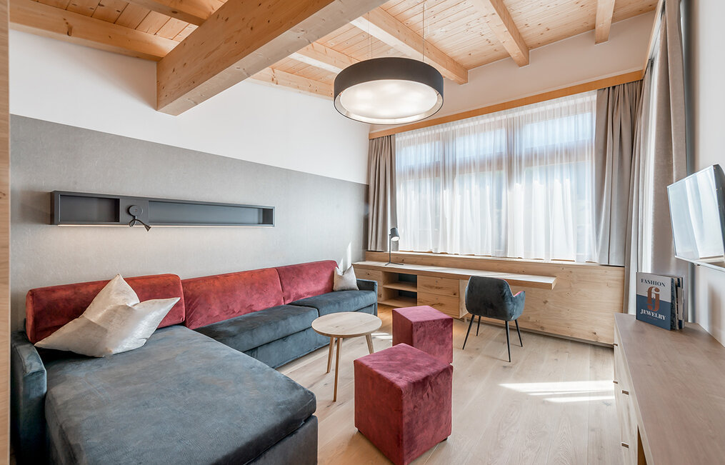A modern living room featuring a dark sofa, red poufs, a round coffee table, a large window with sheer curtains, a wooden desk, a chair, books and a round ceiling light, set in a room with exposed wooden beams.