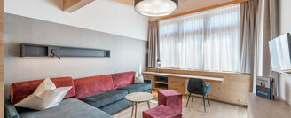 A modern living room featuring a dark sofa, red poufs, a round coffee table, a large window with sheer curtains, a wooden desk, a chair, books and a round ceiling light, set in a room with exposed wooden beams.
