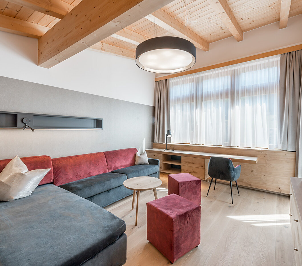 A modern living room featuring a dark sofa, red poufs, a round coffee table, a large window with sheer curtains, a wooden desk, a chair, books and a round ceiling light, set in a room with exposed wooden beams.