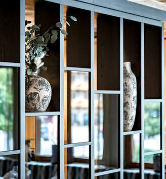 A modern room divider in the form of an open shelving unit featuring decorative vases and green foliage. Warm wooden tones and soft, natural light create a cozy, inviting atmosphere in the interior.