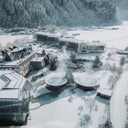 Aerial view of a snow-covered resort with steaming outdoor pools, surrounded by snow-capped trees and buildings with mountains in the background.