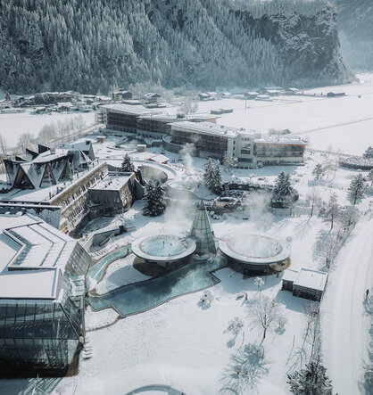 Aerial view of a snow-covered resort with steaming outdoor pools, surrounded by snow-capped trees and buildings with mountains in the background.