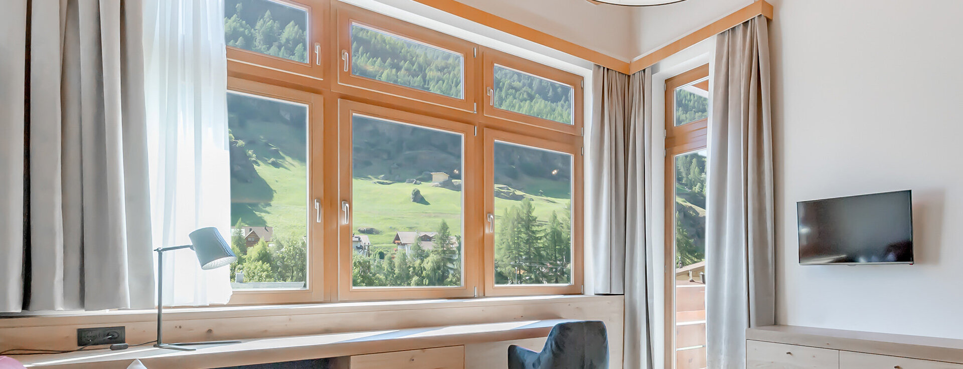 Helles, modernes Wohnzimmer mit großen Fenstern, die einen malerischen Blick auf die Berge bieten, Holzmöbeln, einem an der Wand montierten Fernseher, einem Sofa, zwei Stühlen, einem runden Couchtisch, hellen Vorhängen und viel Tageslicht.