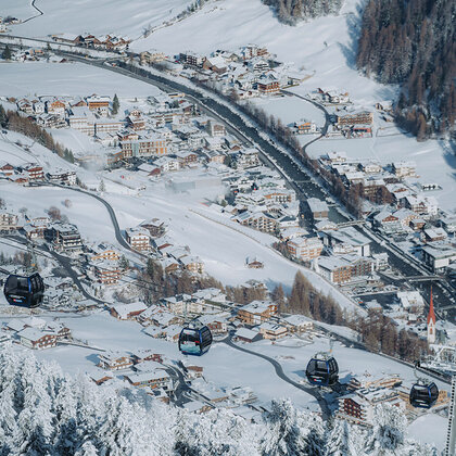 A snow-covered village with houses, winding roads and mountain lifts gliding over the snow-laden trees. The landscape is characterized by pine forests and mountains in the background.