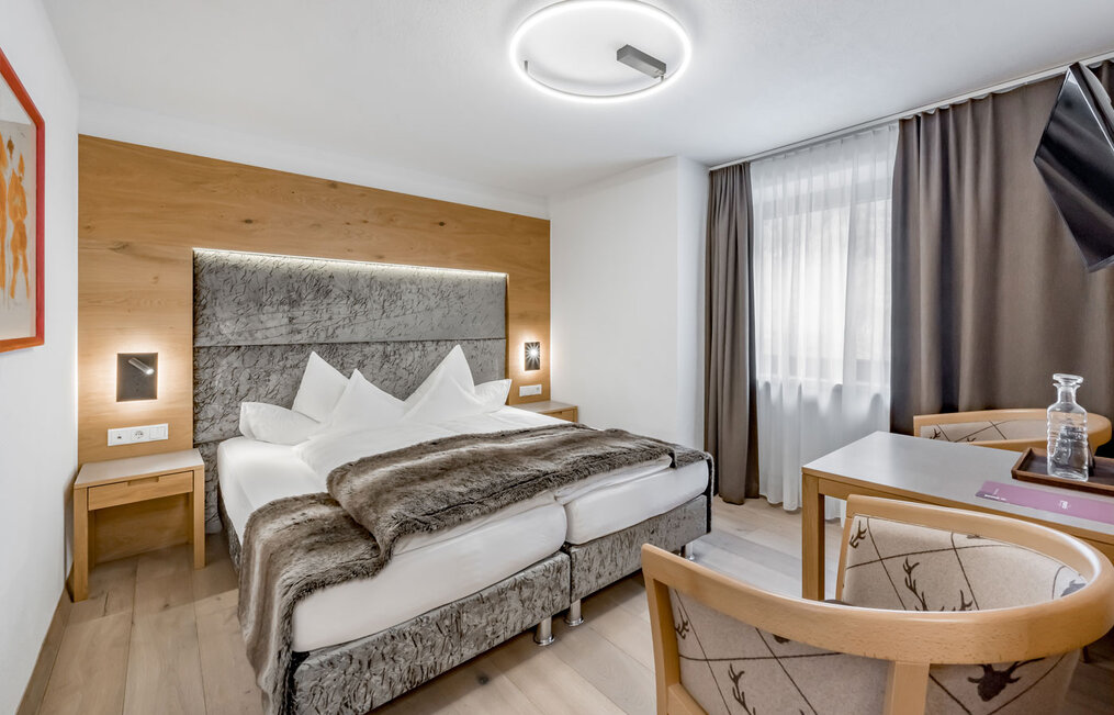 A modern hotel room featuring a double bed, a gray headboard, a faux-fur throw, a small desk and chair, wall art, wood accents and a window with both sheer and opaque curtains. The ceiling light is round and modern.