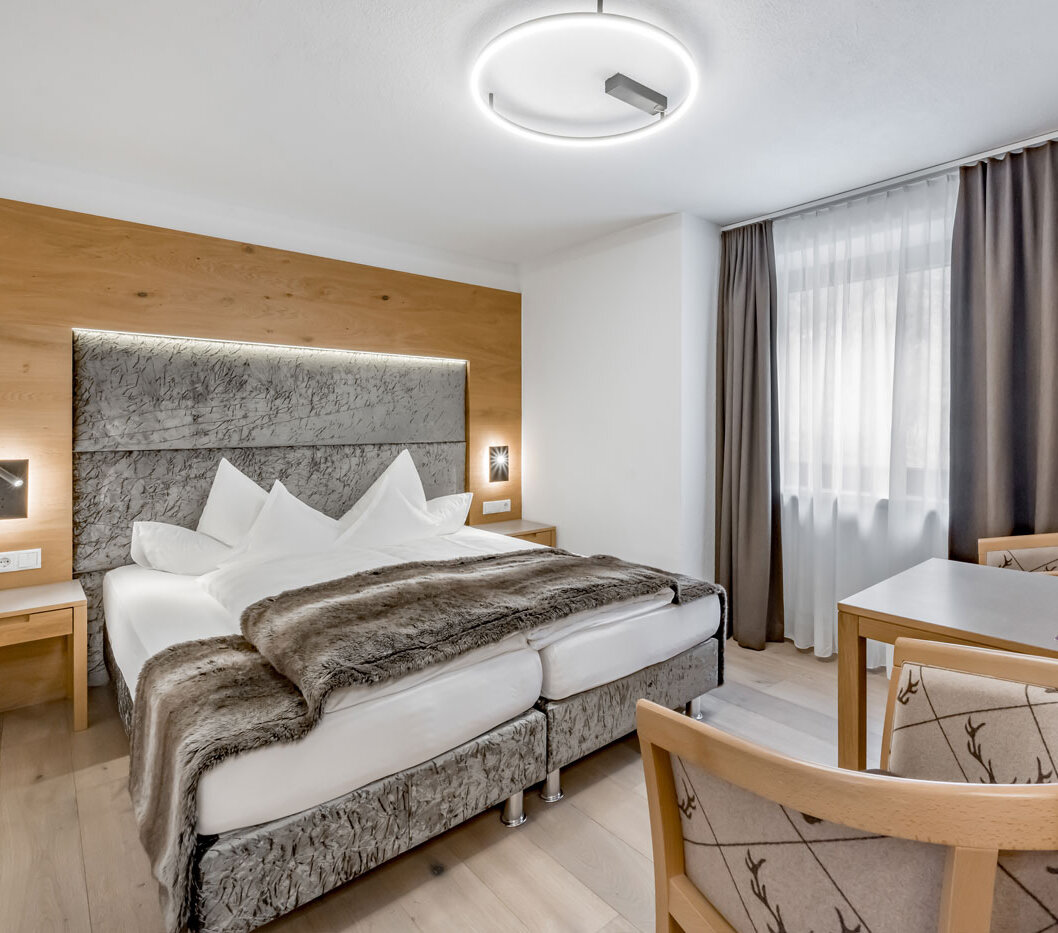 A modern hotel room featuring a double bed, a gray headboard, a faux-fur throw, a small desk and chair, wall art, wood accents and a window with both sheer and opaque curtains. The ceiling light is round and modern.