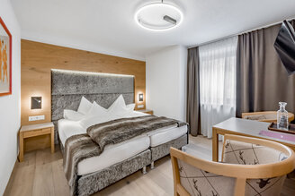 A modern hotel room featuring a double bed, a gray headboard, a faux-fur throw, a small desk and chair, wall art, wood accents and a window with both sheer and opaque curtains. The ceiling light is round and modern.