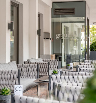 Outdoor seating area with cushioned chairs and small tables, decorated with potted plants. The ambiance is modern and inviting, characterized by neutral tones and glass doors reflecting the surrounding buildings.