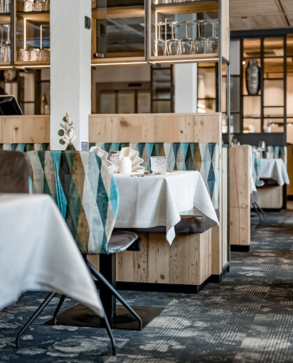 A modern restaurant interior featuring wooden partition elements, patterned chairs, tables set with white tablecloths and neutral décor beneath shelves holding glassware.