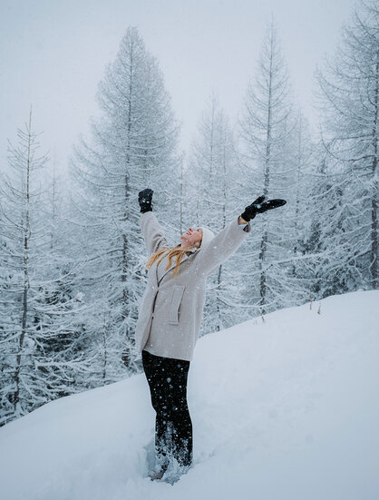 A person stands smiling in deep snow with arms raised, surrounded by snow-covered trees in a winter landscape.