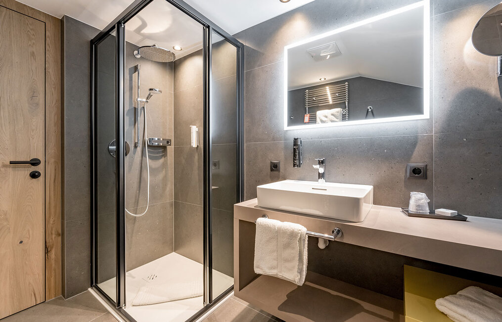 Modern bathroom with a glass shower enclosure, a rainfall shower, an illuminated mirror above a rectangular washbasin and towels neatly arranged on a beige countertop.