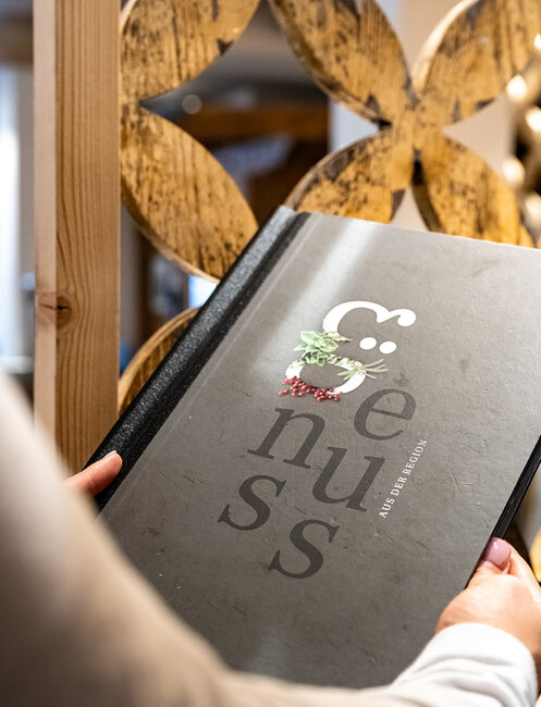 A person is holding a large black menu, the front cover displays the word "Enjoyment" and a small graphic featuring herbs and berries.