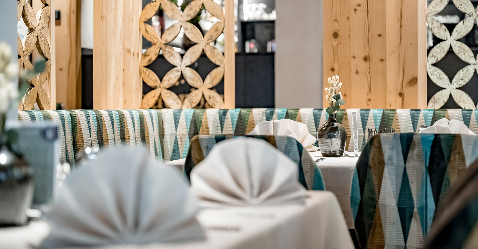 A modern restaurant interior with patterned seating niches, tables with white, folded napkins placed on them and decorative wooden panels with floral cutouts in the background.