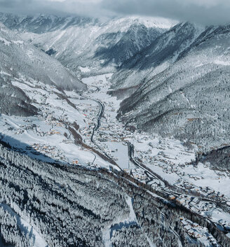 Aerial view of a snow-covered mountain valley with a winding road, scattered buildings, dense pine forests and snow-capped peaks in the background under a cloudy sky.