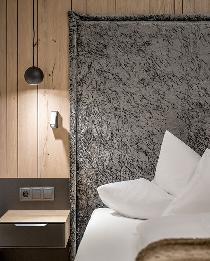 A modern hotel room corner featuring a gray, textured headboard, crisp white pillows, a wooden accent wall, a small nightstand, a wall-mounted light switch and a black pendant light.