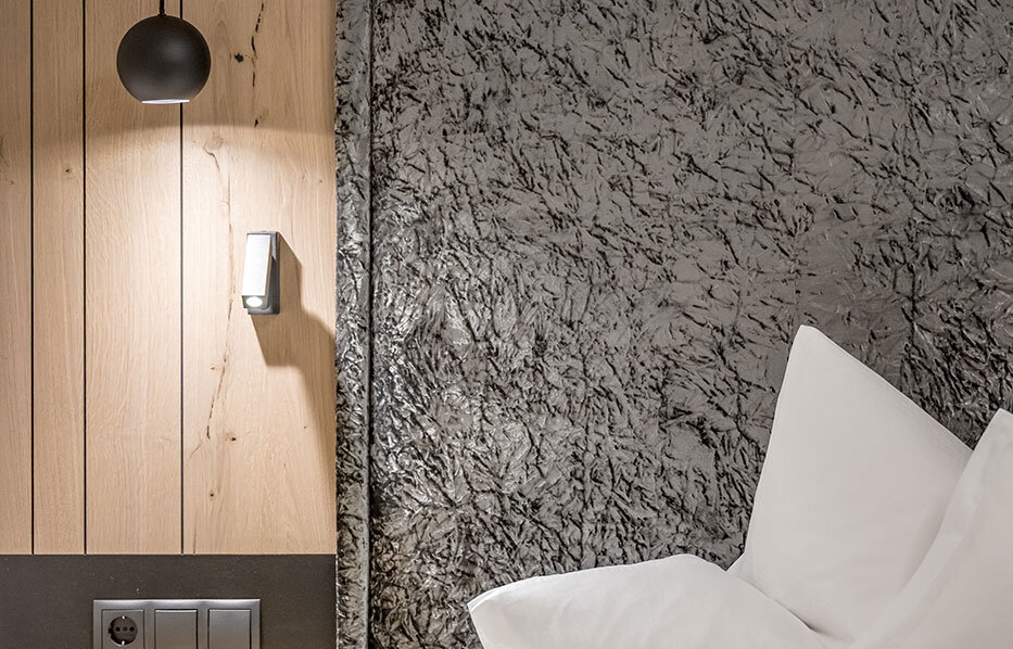 A modern hotel room corner featuring a gray, textured headboard, crisp white pillows, a wooden accent wall, a small nightstand, a wall-mounted light switch and a black pendant light.