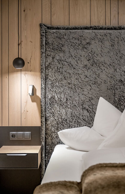 Eine moderne Hotelzimmer-Ecke mit einem grauen, strukturierten Kopfteil, strahlend weißen Kissen, einer Akzentwand aus Holz, einem kleinen Nachttisch, einem an der Wand montierten Lichtschalter und einer schwarzen Pendelleuchte.