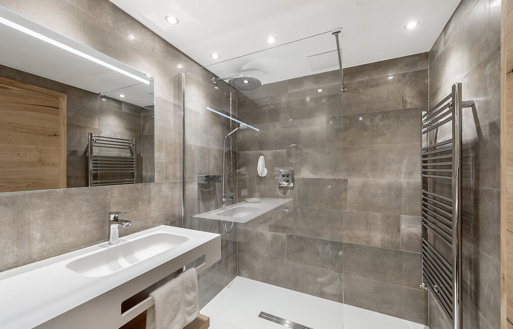 Modernes Badezimmer mit großen grauen Fliesen, einem wandmontierten Waschbecken, einem Handtuchhalter und einer begehbaren Glasdusche. Der Raum ist durch Einbauleuchten gut ausgeleuchtet und verfügt über einen großen Spiegel über dem Waschbecken.