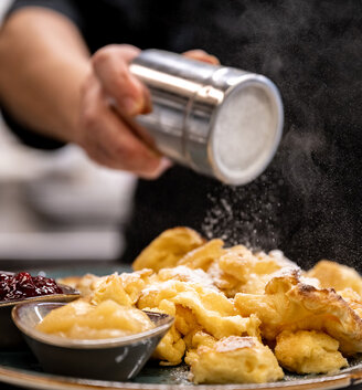Someone is sprinkling powdered sugar onto a plate of Kaiserschmarrn, which is served alongside small bowls of applesauce and red jam.