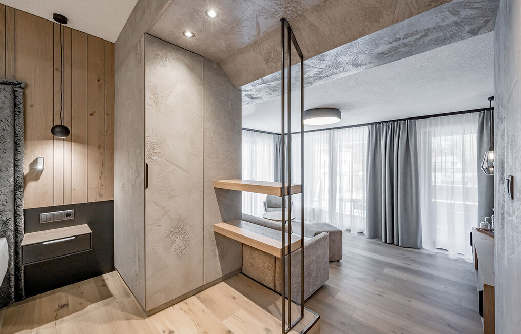 Modern hotel room featuring light wood floors, neutral tones, large windows with sheer and gray curtains, a minimalist bench and a built-in wardrobe. Natural light floods the stylish, spacious room. 