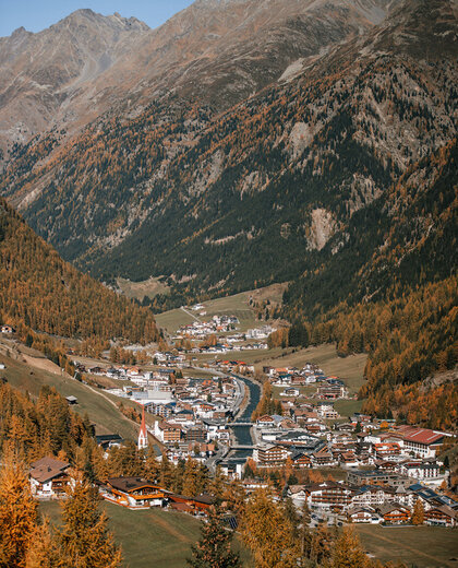 In a green valley, surrounded by trees in autumn colors and high, rugged mountains under a brilliant blue sky, lies a village with houses and a church.