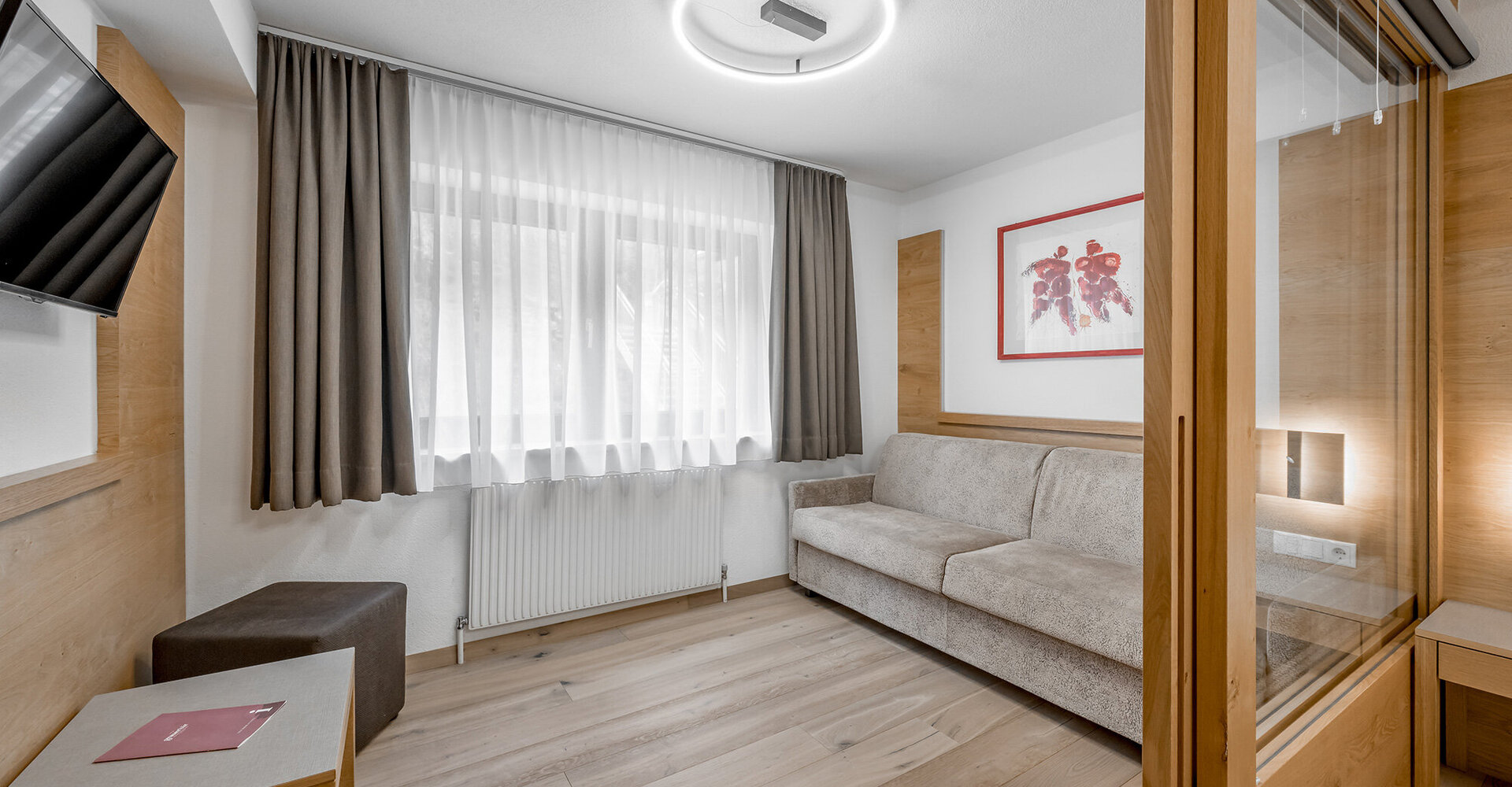 Modernes Wohnzimmer mit einem beigen Sofa, einem an der Wand montierten Fernseher, Holzakzenten, einem Fenster mit transparenten und dunklen Vorhängen, einem kleinen Tisch und einem gerahmte Kunstwerk an der Wand. Der Raum ist hell und von Tageslicht durchflutet.