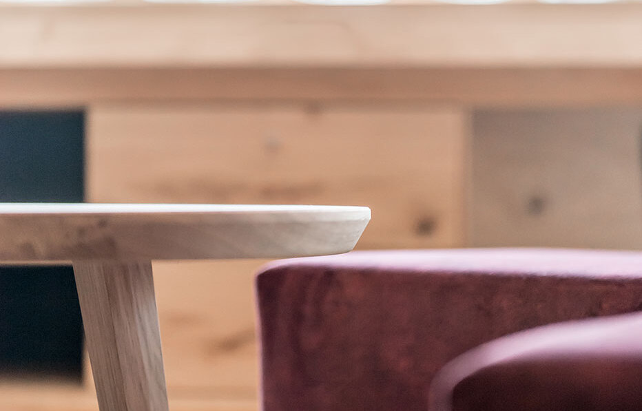 A small wooden table and a burgundy upholstered armchair in a softly lit room, with light streaming in through a large window in the background.