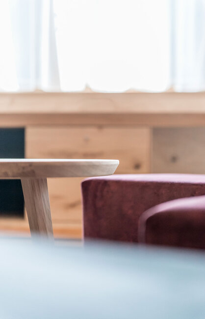 Ein kleiner Holztisch und ein burgunderroter Polstersessel in einem sanft beleuchteten Raum, in den durch ein großes Fenster im Hintergrund Licht hereinströmt.