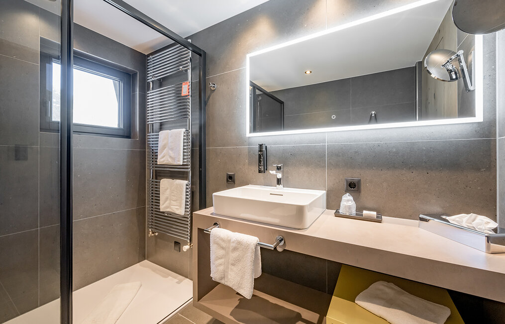 Modern bathroom with a walk-in shower, large illuminated mirror, rectangular sink, gray walls, a towel rack with white towels and a shelf with tissues and toiletries.