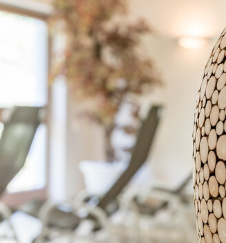 A close-up of a decorative object made of small, round pieces of wood. Blurred deckchairs and a large window are in the background, creating a peaceful atmosphere.