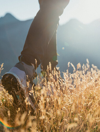 A close-up of a person's legs walking through tall, sunlit grass with blurred mountains in the background while sunlight streams in from the upper right.