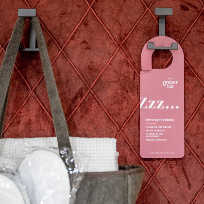 An Haken vor einer rot gesteppten Wand hängen ein rosa Türhänger und eine Tasche mit Handtüchern.