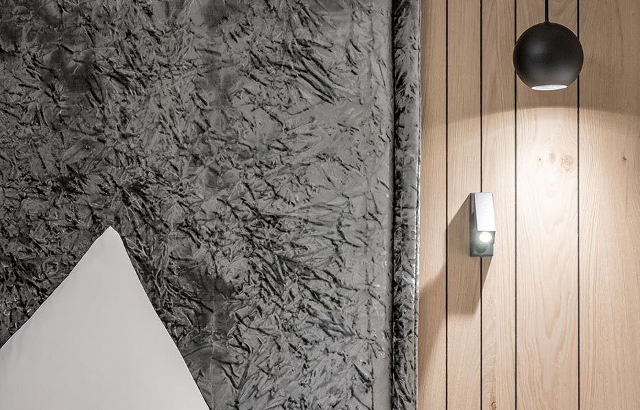 A modern bedroom corner featuring a large, textured gray headboard, white pillows, light wood paneling, a black pendant light, a wall lamp, power outlets and a dark nightstand.