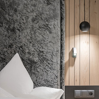 A modern bedroom corner featuring a large, textured gray headboard, white pillows, light wood paneling, a black pendant light, a wall lamp, power outlets and a dark nightstand.