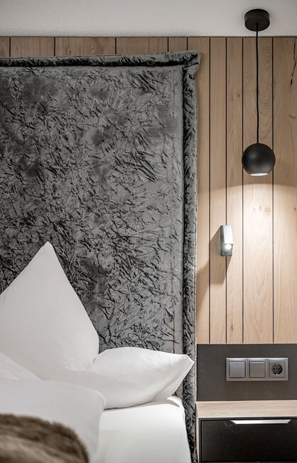 Eine moderne Schlafzimmer-Ecke mit einem großen, grauen Kopfteil mit Struktur, weißen Kissen, hellen Holzvertäfelungen, einer schwarzen Pendelleuchte, einer Wandleuchte, Steckdosen und einem dunklen Nachttisch.