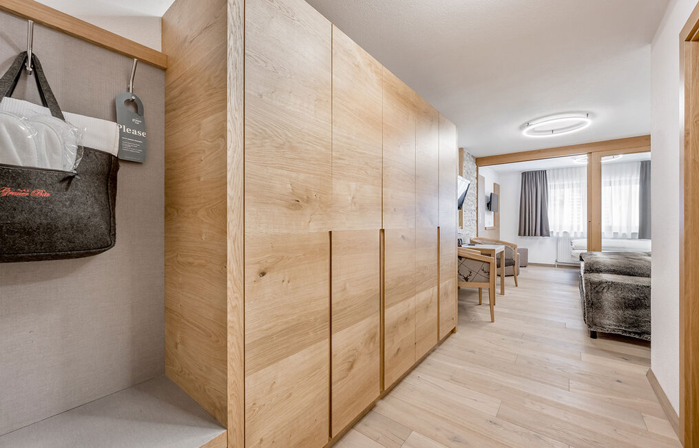 Modernes Hotelzimmer mit hellen Holzböden und Schränken, einer Sitzecke mit Stühlen und einem Sofa, großen Fenstern mit transparenten Vorhängen sowie einem an der Wand montierten Fernseher. Auf der linken Seite in der Nähe des Eingangs hängt eine schwarze Filztasche.
