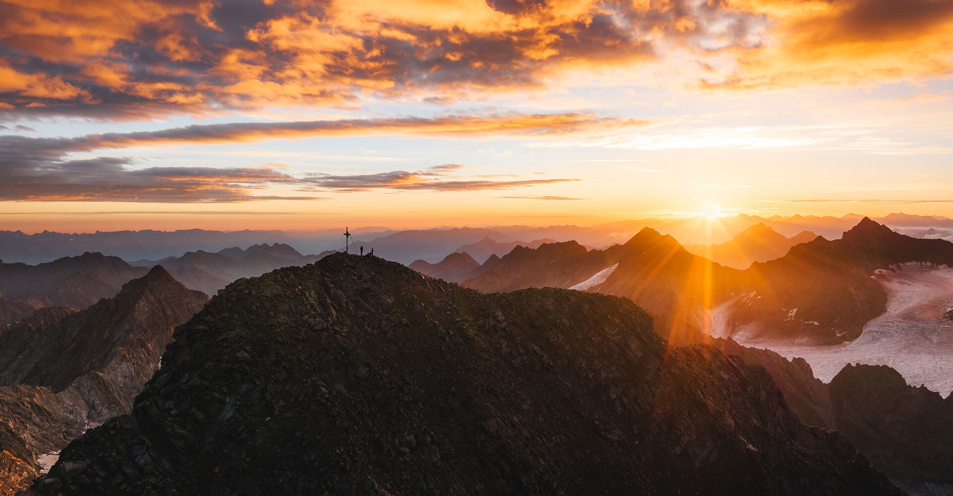 A sunset over rugged mountain peaks with a radiant sunbeam and colorful clouds. On the summit stands a small cross with its silhouette against the luminous sky.