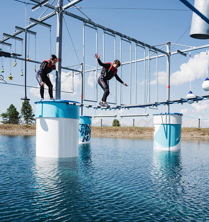 Zwei Personen in Klettergurten balancieren auf einer hängenden Seilbrücke über dem Wasser und bewältigen unter strahlend blauem Himmel einen Outdoor-Abenteuer-Hindernisparcours.
